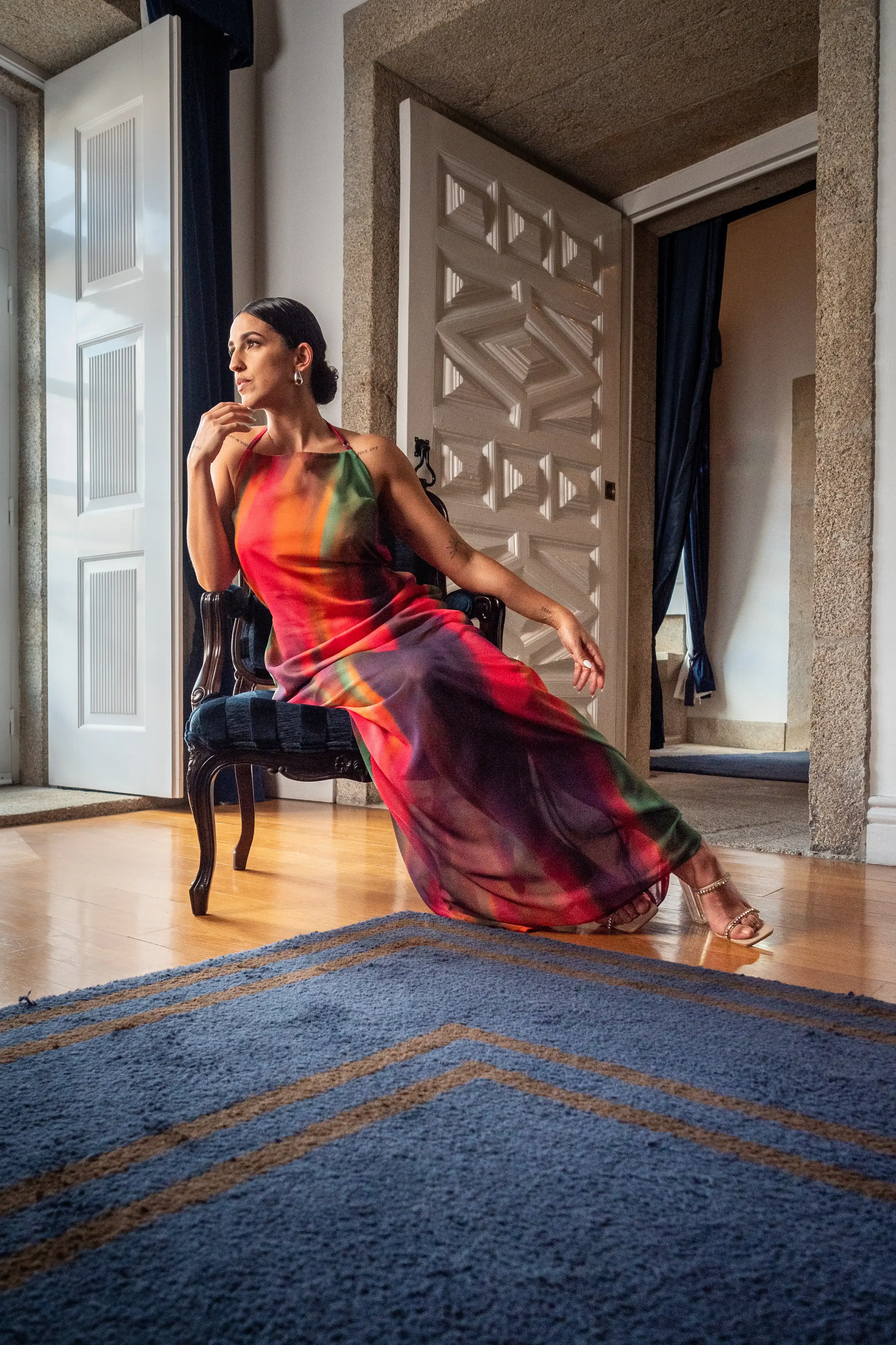 Mulher posa com um vestido comprido de uma cores vivas sentada na cadeira de uma sala requintada.